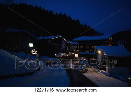 Stock Photograph - Snow covered rooves in the Alpine village at night with snowflake light and Christmas decorations; Filzmoos, Austria. Fotosearch