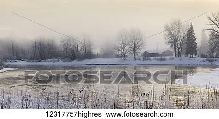 Stock Photograph - Soft golden sky and a tranquil lake at sunrise in winter; Prince George, British Columbia, Canada. Fotosearch