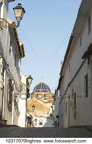 Stock Photograph - Street in downtown Altea, a beautiful town in Costa Blanca, with the cupule of the church in the background; Altea, Alicante, Spain. Fotosearch