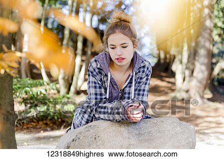 Stock Photograph - This young teenage girl hangs out alone in a park, sitting on a rock in a thoughtful disengaged position thinking to herself; New Westminster, British Columbia, Canada. Fotosearch