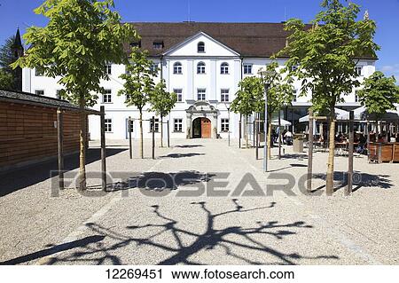 Stock Image - Trees lining a walkway to a building and restaurant customers sitting on an outdoor patio; Die Herreninsel im Chiemsee, Bayern, Germany. Fotosearch