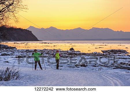 Stock Image - Two back lit people cross country skiing on the Tony Knowles Coastal Trail at sunset, Southcentral Alaska. Fotosearch