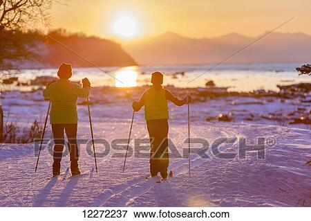 Stock Photo - Two back lit people cross country skiing on the Tony Knowles Coastal Trail at sunset, Southcentral Alaska. Fotosearch