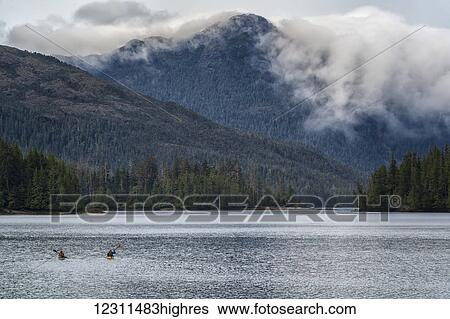 Stock Photograph - Two kayakers paddling toward the mainland while clouds flow over the coastal mountains; Haida Gwaii, British Columbia, Canada. Fotosearch
