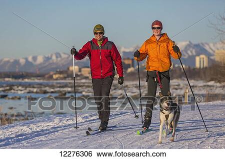Stock Photograph - Two people cross country ski and skijor with an Alaska Husky on the Tony Knowles Coastal Trail near Earthquake Park with Anchorage skyline in the background, Cook Inlet, Southcentral Alaska. Fotosearch