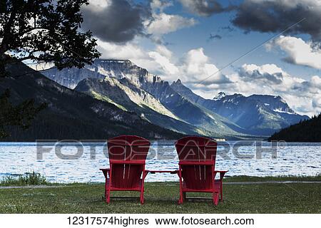 Stock Photograph - Two red adirondack chairs sit facing the lake and Rocky mountains, Waterton Lakes National Park; Alberta, Canada. Fotosearch
