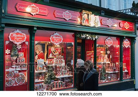 Stock Image - Two women walk past a gift shop, Reber, selling Mozart-branded Mozartkugeln chocolates, truffles and marzipan sweets; Salzburg, Austria. Fotosearch