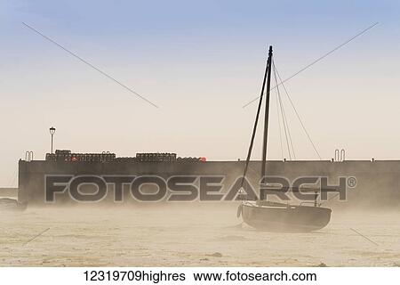 Stock Photograph - Wind blows sand up on the beach against a sailboat sitting on the shore; Lindisfarne, Northumberland, England. Fotosearch