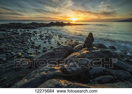 Picture - Woman sits on the shore of the Pacific Ocean watching the sunset near Jordan River; Vancouver Island, British Columbia, Canada. Fotosearch