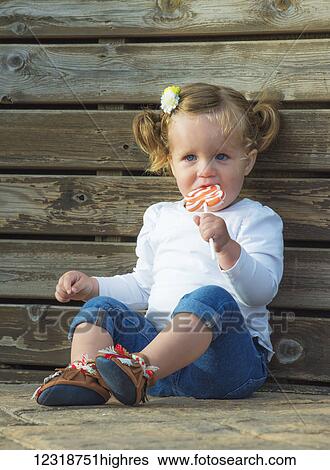 Stock Photograph - Young girl with blond hair and blue eyes sitting and eating a lollipop; Tarifa, Cadiz, Andalusia, Spain. Fotosearch