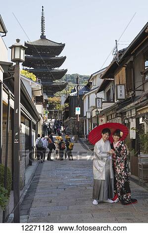 Stock Photo - Young Japanese couple in kimono posing under a red umbrella in a narrow street with pagoda in the distance; Kyoto, Japan. Fotosearch