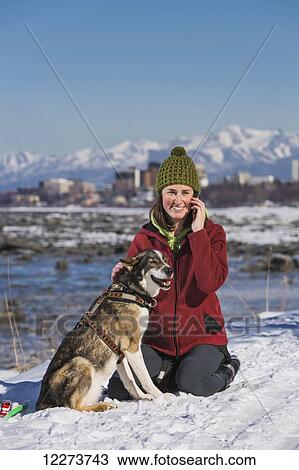 Stock Image - Young woman with her Alaska Husky talks on a cell phone along the Tony Knowles Coastal Trail, Anchorage, Southcentral Alaska. Fotosearch