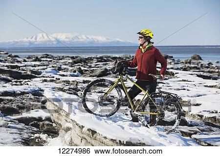 Stock Photograph - Young woman with her bicycle stands on frozen ice chunks along the Tony Knowles Coastal Trail, Anchorage, Southcentral Alaska. Fotosearch