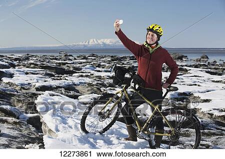 Stock Image - Young woman with her bicycle takes a selfie with a smart phone while standing on frozen ice chunks along the Tony Knowles Coastal Trail, Anchorage, Southcentral Alaska. Fotosearch