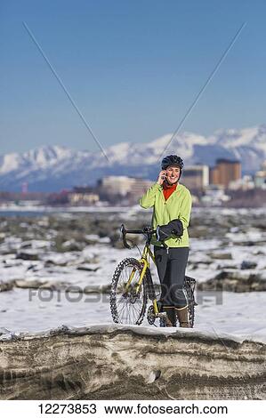 Stock Image - Young woman with her bicycle talks on a cell phone while standing on frozen ice chunks along the Tony Knowles Coastal Trail, Anchorage, Southcentral Alaska. Fotosearch