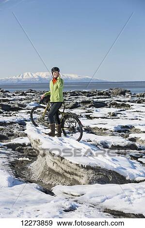 Stock Photo - Young woman with her bicycle talks on a cell phone while standing on frozen ice chunks along the Tony Knowles Coastal Trail, Anchorage, Southcentral Alaska. Fotosearch