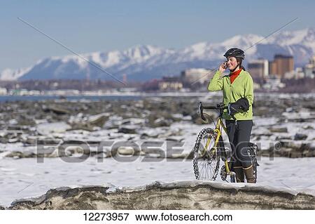 Stock Photo - Young woman with her bicycle talks on a cell phone while standing on frozen ice chunks along the Tony Knowles Coastal Trail, Anchorage, Southcentral Alaska. Fotosearch