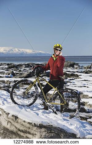 Stock Photo - Young woman with her bicycle talks on a cell phone while standing on frozen ice chunks along the Tony Knowles Coastal Trail, Anchorage, Southcentral Alaska. Fotosearch