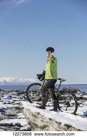 Stock Photograph - Young woman with her bicycle talks on a cell phone while standing on frozen ice chunks along the Tony Knowles Coastal Trail, Anchorage, Southcentral Alaska. Fotosearch