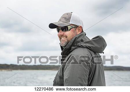 Stock Photograph - A man wearing sunglasses and a hat stands on a fishing boat with a view of the water and coastline; Maine, United States of America. Fotosearch