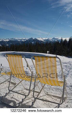 Stock Photograph - A pair of yellow and white striped deck chairs for customers in the sunshine outside a cafe at the top of a ski piste with views across the mountain peaks of the Austrian Alps; Filzmoos, Austria. Fotosearch