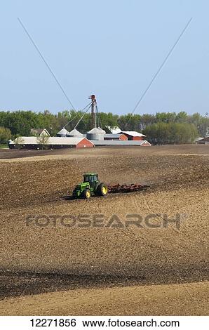 Stock Photograph - A tractor and field implement prepare a field for spring planting with a swine confinement facility in the background, near Truman; Minnesota, United States of America. Fotosearch