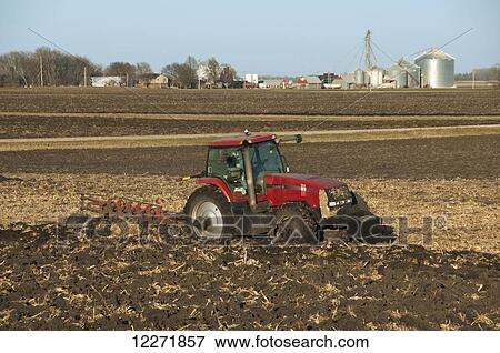 Stock Photo - A tractor pulling a moldboard plough prepares a field for spring planting with a farmstead in the background, near Truman; Minnesota, United States of America. Fotosearch