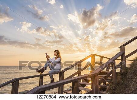 Picture - A woman sits on a wooden railing at the water's edge using a cell phone; Tarifa, Cadiz, Andalusia, Spain. Fotosearch