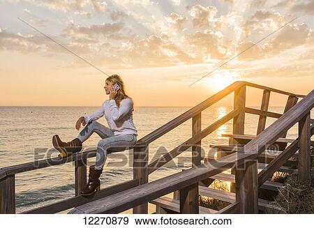Stock Photo - A woman sits on a wooden railing at the water's edge using a cell phone; Tarifa, Cadiz, Andalusia, Spain. Fotosearch