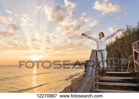 Stock Photo - A young woman stands on wooden steps at the water's edge looking up with arms raised; Tarifa, Cadiz, Andalusia, Spain. Fotosearch