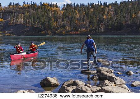 Stock Photograph - Family on North Saskatchewan River, adults canoeing and son playing on the rocks; Edmonton, Alberta, Canada. Fotosearch