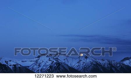 Stock Image - Full moon in behind the rocky mountains in winter, Banff National Park; Banff, Alberta, Canada. Fotosearch
