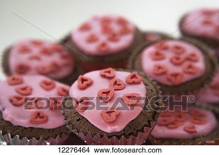 Picture - Homemade chocolate fairy cakes in gold and silver cases with pink icing and red heart-shaped sprinkles against a white background; London, England. Fotosearch