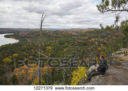 Stock Photo - Man sitting on the edge of a cliff atop Booth's Rock with a view of Rock Lake, Algonquin Park; Ontario, Canada. Fotosearch