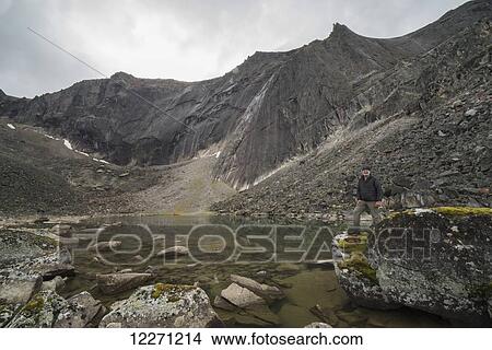 Man standing on a rock beside an unnamed lake in the Tombstone Mountains; Yukon Territory, Canada View Large Photo Image Picture - Man standing on a rock beside an unnamed lake in the Tombstone Mountains; Yukon Territory, Canada. Fotosearch