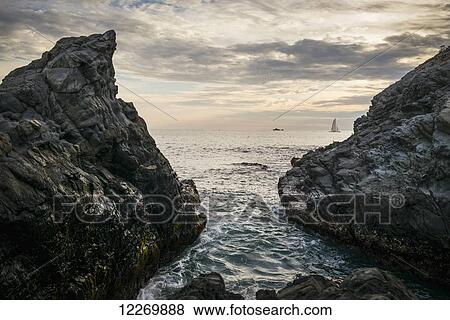 Stock Photo - Rugged rocks on the coast with a view of ship and sailboat in the distance at sunset; Ixtapa-Zihuatanejo, Guerrero, Mexico. Fotosearch