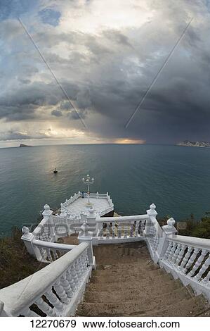 Stock Photo - Steps leading down to the turquoise water with the sun glowing on the horizon under storm clouds; Benidorm, Spain. Fotosearch