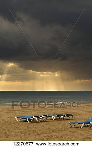 Sun rays shine through the dark clouds over the ocean and lounge chairs on the beach; Benidorm, Spain View Large Photo Image Stock Photo - Sun rays shine through the dark clouds over the ocean and lounge chairs on the beach; Benidorm, Spain. Fotosearch