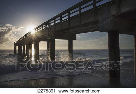 Stock Image - Sunlight at sunrise streaming through the rails of an ocean pier as waves crash on the pier pylons; United States of America. Fotosearch