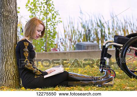 Stock Image - Young disabled woman sitting beside her wheelchair reading a book in a city park in autumn; Edmonton, Alberta, Canada. Fotosearch