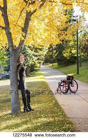 Stock Photo - Young paraplegic woman standing against a tree without her wheelchair on a path in a city park in autumn; Edmonton, Alberta, Canada. Fotosearch