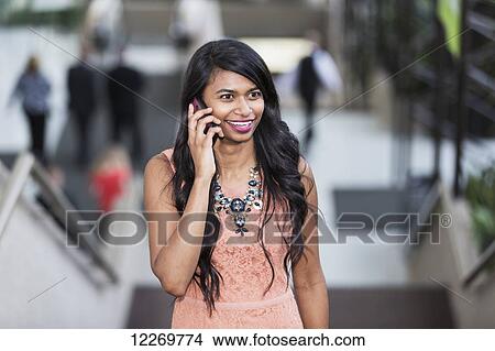 Picture - Young professional businesswoman talking on her smart phone on escalator of office building; Edmonton, Alberta, Canada. Fotosearch