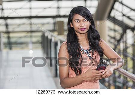 Stock Image - Young professional businesswoman holding a smart phone in lobby of office building; Edmonton, Alberta, Canada. Fotosearch