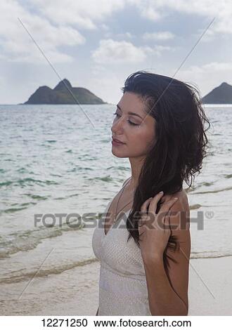 Stock Image - Young woman in white dress on the beach at the water's edge; Kailua, Island of Hawaii, Hawaii, United States of America. Fotosearch