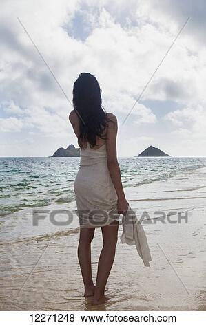Stock Photo - Young woman in white dress on the beach at the water's edge; Kailua, Island of Hawaii, Hawaii, United States of America. Fotosearch