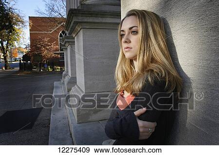Stock Photo - Young woman leaning against a stone pillar with her arms crossed in front of her, looking unhappy; United States of America. Fotosearch