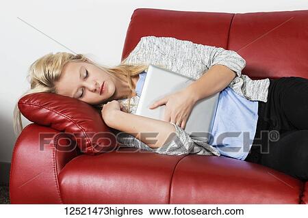 Stock Photograph - A beautiful young millennial business woman catching a nap on a red couch in a workplace and holding her tablet; Sherwood Park, Alberta, Canada. Fotosearch