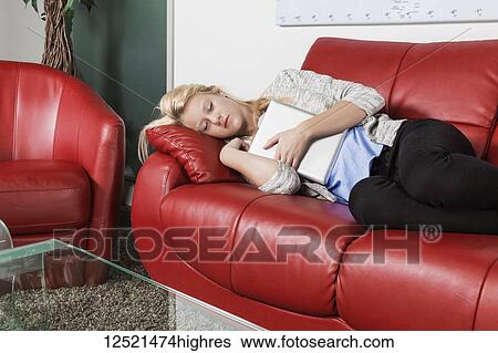 Stock Photograph - A beautiful young millennial business woman catching a nap on a red couch in a workplace and holding her tablet; Sherwood Park, Alberta, Canada. Fotosearch