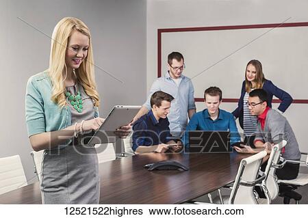 Stock Photograph - A beautiful young millennial business woman working on her tablet in a conference room with her co-workers; Sherwood Park, Alberta, Canada. Fotosearch