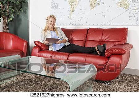 Stock Photograph - A beautiful young millennial business woman with long blond hair relaxing in the lobby of a workplace and using her tablet; Sherwood Park, Alberta, Canada. Fotosearch